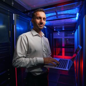 Bearded smiling system administrator is working with server rack and telecommunication equipments in