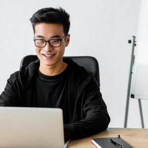 smiling seo manager in glasses sitting at table and using laptop